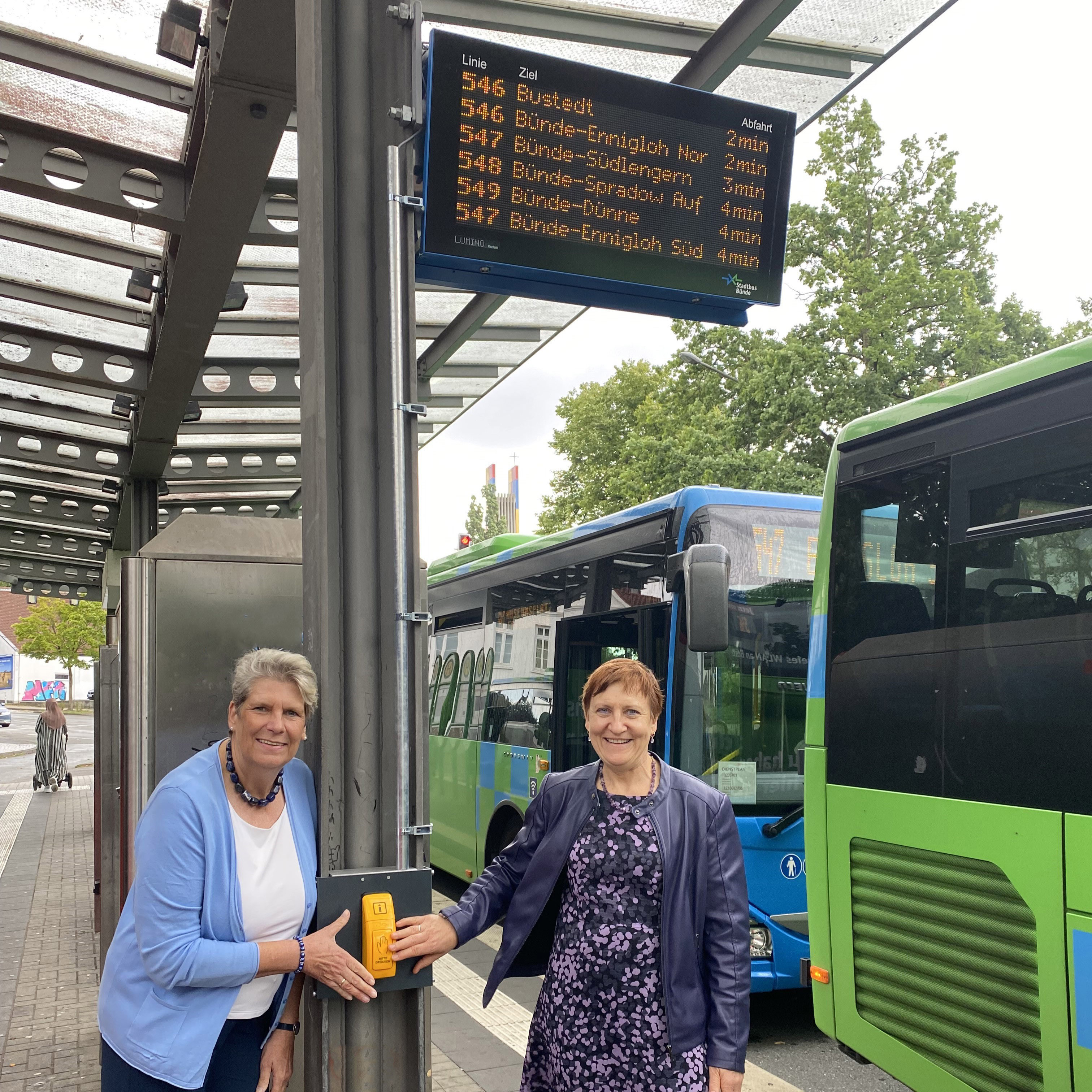 Bürgermeisterin Susanne Rutenkröger und Geschäftsführerin Dr. Marion Kapsa zeigen die digitale Anzeigetafel an der Haltestelle